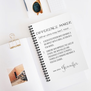 May include: A notebook with a black spiral binding and a white cover. The cover has the text "DIFFERENCE MAKER" in black with the phonetic pronunciation in brackets. The text "See also: Jennifer" is written in black at the bottom of the page.