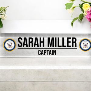 May include: A clear glass desk nameplate with a black border and the text "SARAH MILLER CAPTAIN" in black lettering. The nameplate has two gold and blue United States Navy seals on either side of the text.