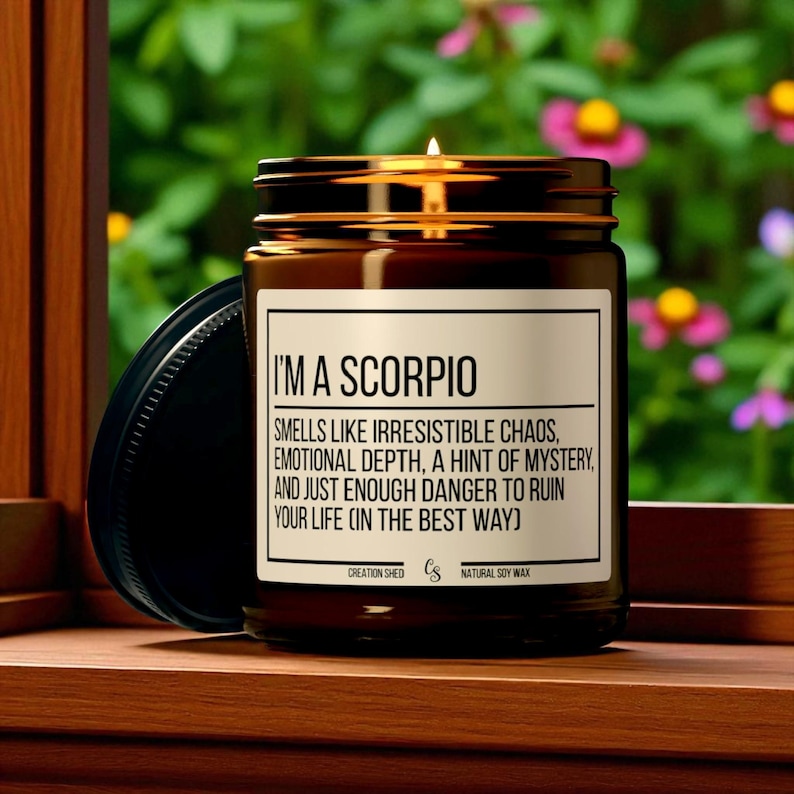 Peut inclure: Une bougie en pot de verre brun allum&eacute;e avec un couvercle noir. La bougie porte une &eacute;tiquette indiquant "I'M A SCORPIO" et d&eacute;crit le parfum. La bougie est sur une surface en bois avec un arri&egrave;re-plan floral vert et rose flou.