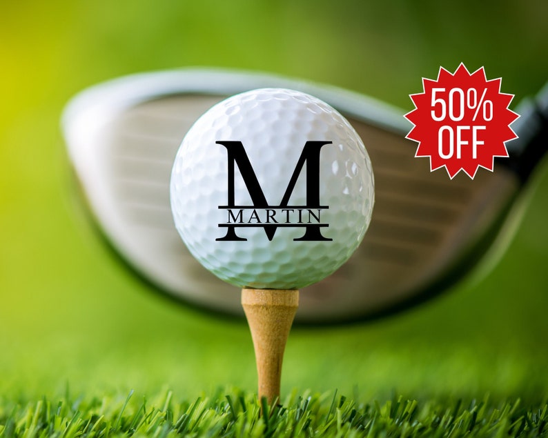 May include: A white golf ball with a black letter "M" and the name "MARTIN" printed on it. The golf ball is on a tee and a golf club is in the background.