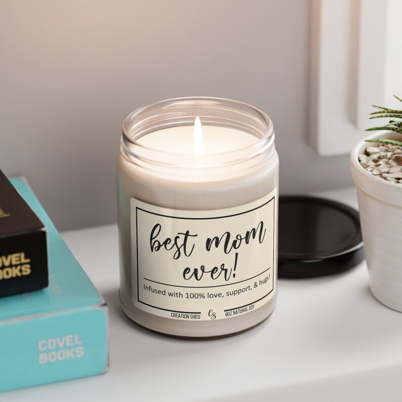 K&ouml;nnte beinhalten: Eine brennende, beige Kerze in einem Glas mit schwarzem Deckel. Auf dem Etikett steht "best mom ever!" mit dem Text "Infused with 100% love, support, & hugs!". Die Kerze ist 9 oz und besteht aus nat&uuml;rlichem Soja.