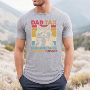 K&ouml;nnte beinhalten: Graues T-Shirt mit Retro-Design, das einen Waschb&auml;ren mit Sonnenbrille zeigt, der ein Getr&auml;nk und Pommes h&auml;lt, mit dem Text "DAD TAX MAKING SURE IT'S NOT POISON". Ein l&auml;ssiges Kleidungsst&uuml;ck.