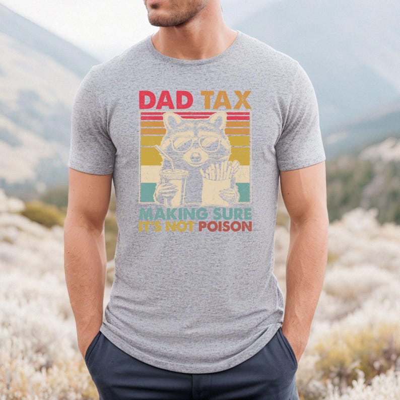 K&ouml;nnte beinhalten: Graues T-Shirt mit Retro-Design, das einen Waschb&auml;ren mit Sonnenbrille zeigt, der ein Getr&auml;nk und Pommes h&auml;lt, mit dem Text "DAD TAX MAKING SURE IT'S NOT POISON". Ein l&auml;ssiges Kleidungsst&uuml;ck.