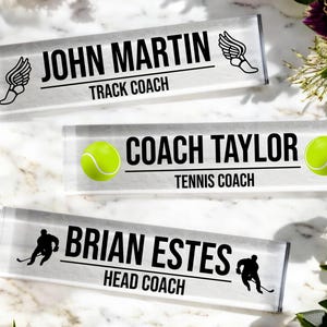 K&ouml;nnte beinhalten: Drei transparente Acryl-Schreibtisch-Namensschilder mit schwarzem Text und Grafiken. Das obere Namensschild lautet "John Martin Track Coach" mit einer Grafik eines Laufschuhs. Das mittlere Namensschild lautet "Coach Taylor Tennis Coach" mit einer Grafik eines Tennisballs. Das untere Namensschild lautet "Brian Estes Head Coach" mit einer Grafik eines Hockeyspielers.