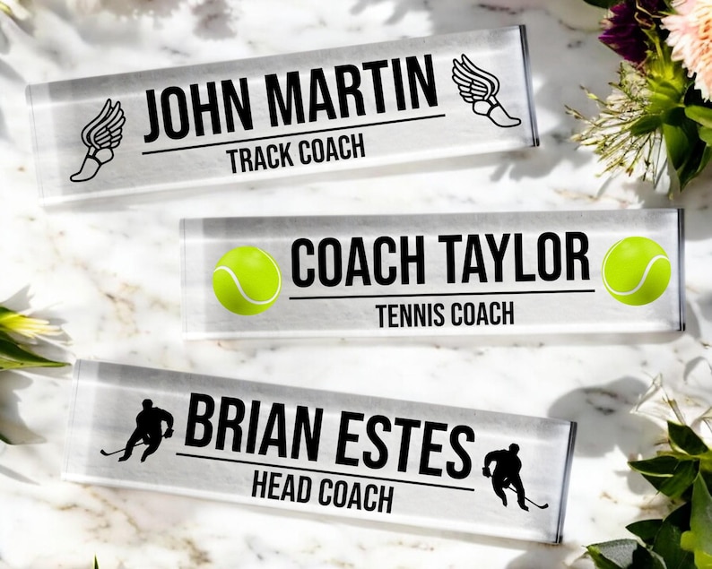 K&ouml;nnte beinhalten: Drei transparente Acryl-Schreibtisch-Namensschilder mit schwarzem Text und Grafiken. Das obere Namensschild lautet "John Martin Track Coach" mit einer Grafik eines Laufschuhs. Das mittlere Namensschild lautet "Coach Taylor Tennis Coach" mit einer Grafik eines Tennisballs. Das untere Namensschild lautet "Brian Estes Head Coach" mit einer Grafik eines Hockeyspielers.