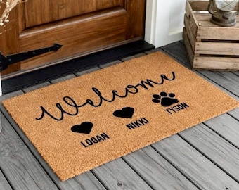 May include: A rectangular coir doormat with the word "Welcome" in a cursive font. Below the word are three black graphics: a heart, a heart, and a paw print. The names "Logan", "Nikki", and "Tyson" are below the graphics.
