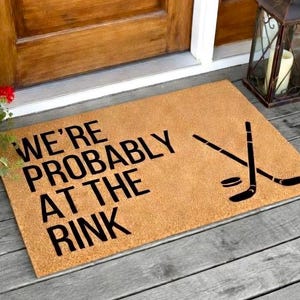 May include: A rectangular doormat made of a tan material with the text "WE'RE PROBABLY AT THE RINK" in black. The mat also features a black hockey puck and crossed hockey sticks. The mat is placed on a wooden porch.