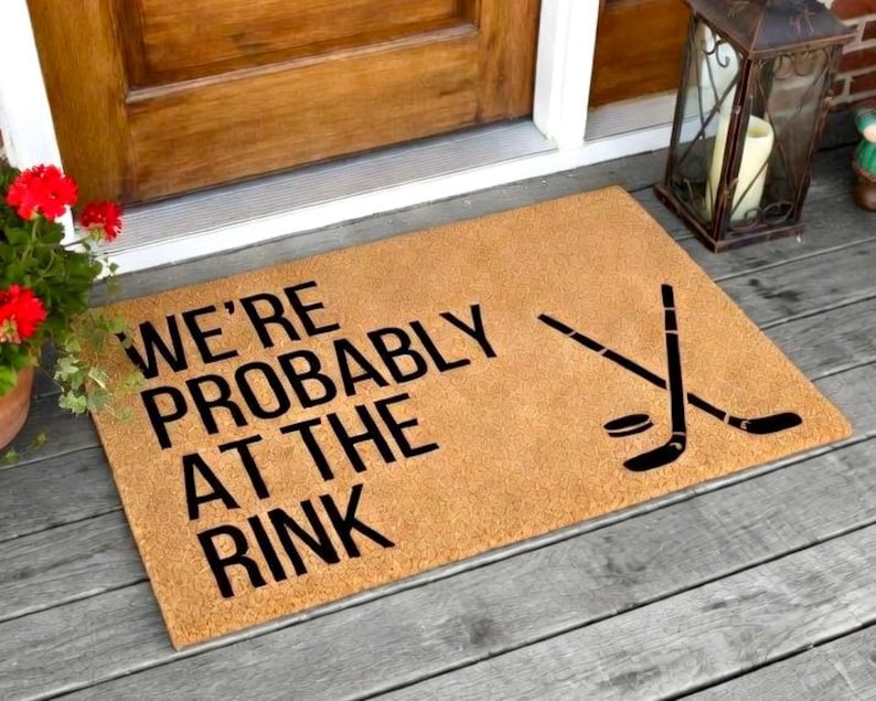May include: A rectangular doormat made of a tan material with the text "WE'RE PROBABLY AT THE RINK" in black. The mat also features a black hockey puck and crossed hockey sticks. The mat is placed on a wooden porch.