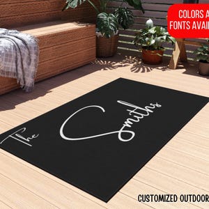 Puede incluir: Alfombra de exterior negra con el texto "The Smiths" en fuente cursiva blanca. La alfombra es personalizable con diferentes colores y fuentes.