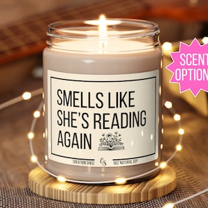 May include: A brown soy candle with a white label that reads "Smells Like She's Reading Again". The label also includes a small illustration of a book with flowers. The candle is on a wooden coaster and surrounded by white fairy lights.