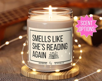 May include: A brown soy candle with a white label that reads "Smells Like She's Reading Again". The label also includes a small illustration of a book with flowers. The candle is on a wooden coaster and surrounded by white fairy lights.