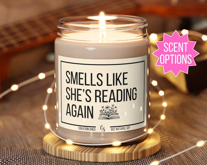 May include: A brown soy candle with a white label that reads "Smells Like She's Reading Again". The label also includes a small illustration of a book with flowers. The candle is on a wooden coaster and surrounded by white fairy lights.