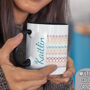 May include: White ceramic mug with a black handle and interior. The mug features the name "Kaitlin" in teal script on one side and a colorful geometric pattern on the other. The mug is held by a person. The text "MULTIPLE COLOR OPTIONS" is also visible.