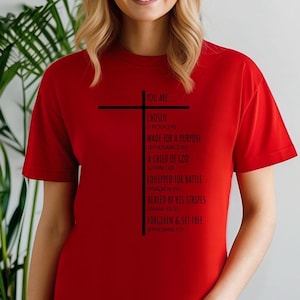 May include: Red t-shirt with a black cross and the text "You are... Chosen (1 Peter 2:9) Made for a Purpose (Ephesians 2:10) A Child of God (John 1:12) Equipped for Battle (Psalm 18:39) Healed by His Stripes (Isaiah 53:5) Forgiven & Set Free (Ephesians 1:7)"
