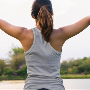 May include: A woman wearing a gray tank top stands with her arms outstretched, facing away from the camera. She is in a park setting with green trees and a body of water in the background.