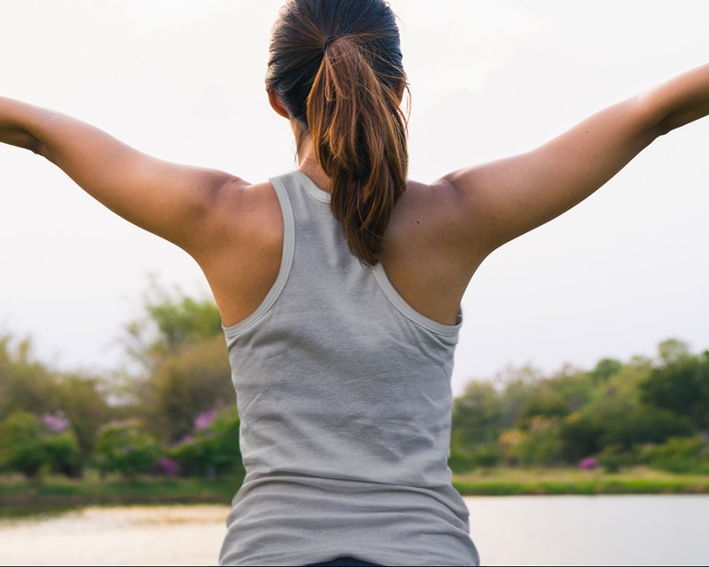 May include: A woman wearing a gray tank top stands with her arms outstretched, facing away from the camera. She is in a park setting with green trees and a body of water in the background.