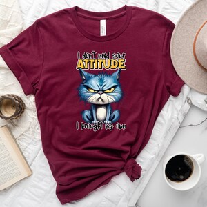Op de afbeelding: Een maroon t-shirt met een cartoonkat met een chagrijnige uitdrukking. De tekst op het shirt luidt "I don't need your attitude I brought my own".