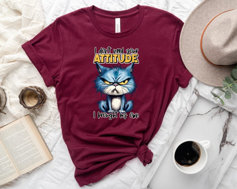 Op de afbeelding: Een maroon t-shirt met een cartoonkat met een chagrijnige uitdrukking. De tekst op het shirt luidt "I don't need your attitude I brought my own".