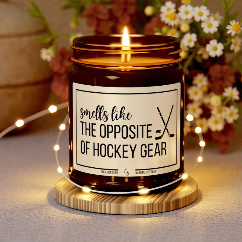 May include: A lit candle in a brown glass jar with a white label that reads "smells like the opposite of hockey gear." The candle sits on a wooden coaster and is surrounded by fairy lights and flowers.