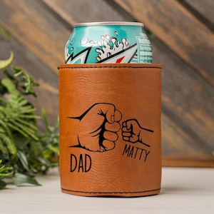 May include: Brown leather can cooler with a black design of two fists bumping. The text "DAD" is under the larger fist and "MATTY" is under the smaller fist.
