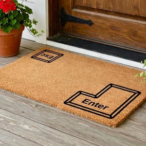May include: A rectangular doormat made of natural fibers, featuring a black outline of the "Enter" key and the word "Enter" in the center. The "ESC" key is also outlined in black. The mat is placed on a wooden porch.