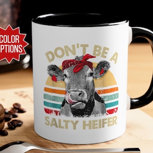 May include: White ceramic mug with a black rim and handle. The mug features a retro-style illustration of a cow with a red bandana, and the text "Don't be a Salty Heifer".