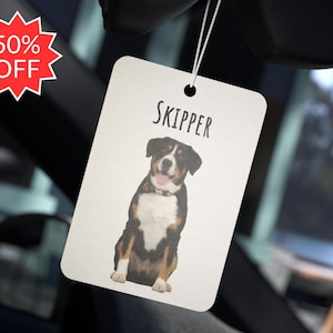May include: White car air freshener with a brown and white dog on it. The dog is sitting and has the name "SKIPPER" printed above it.