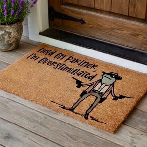 May include: A rectangular doormat made of natural fibers. The mat features a cartoon frog wearing a cowboy hat and holding two guns. Text on the mat reads "Hold on Partner, I'm Overstimulated."