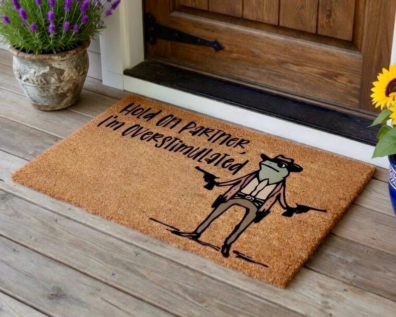 May include: A rectangular doormat made of natural fibers. The mat features a cartoon frog wearing a cowboy hat and holding two guns. Text on the mat reads "Hold on Partner, I'm Overstimulated."