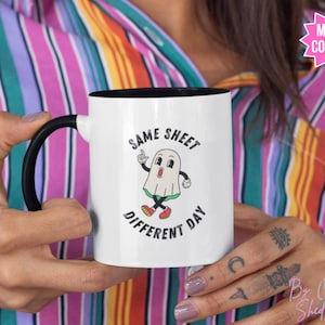 Op de afbeelding: Witte keramische mok met een zwarte handgreep en zwarte rand. De mok heeft een cartoon-geest met de tekst "Same Sheet Different Day" erop gedrukt.
