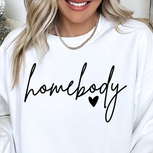 K&ouml;nnte beinhalten: Wei&szlig;er Pullover mit dem Text "homebody" in schwarzer Schrift mit einem kleinen schwarzen Herzen.