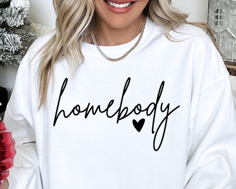 K&ouml;nnte beinhalten: Wei&szlig;er Pullover mit dem Text "homebody" in schwarzer Schrift mit einem kleinen schwarzen Herzen.