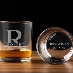 May include: Two clear whiskey glasses, one upright and filled with amber liquid, the other inverted. The upright glass is engraved with the letter "R" over the name "ROBERT". The inverted glass reads "GROOMSMAN 09.15.24".