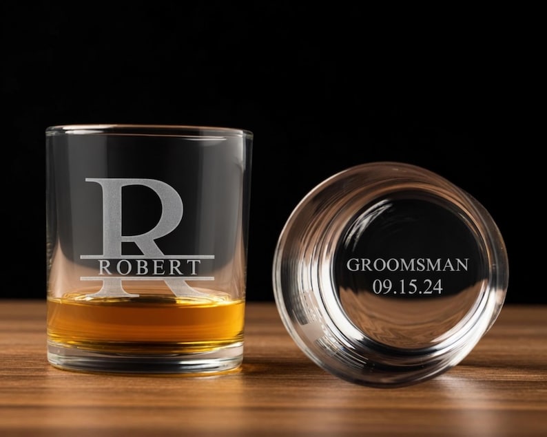 May include: Two clear whiskey glasses, one upright and filled with amber liquid, the other inverted. The upright glass is engraved with the letter "R" over the name "ROBERT". The inverted glass reads "GROOMSMAN 09.15.24".