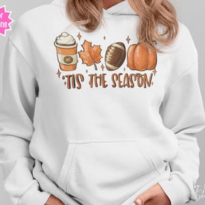 Op de afbeelding: Witte hoodie met een herfstthema-afbeelding. Het ontwerp bevat een pumpkin spice latte, een esdoornblad, een voetbal en een pompoen. De zin "'Tis the Season" staat onder de afbeeldingen. De hoodie heeft een voorzak en een capuchon met trekkoord.