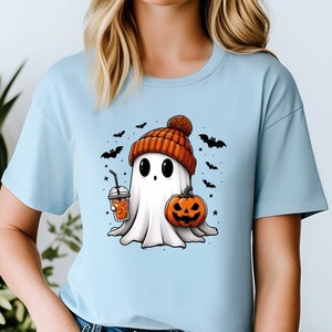 May include: A light blue t-shirt featuring a cartoon ghost wearing a knitted hat and holding a pumpkin and a cup of orange liquid. The ghost is surrounded by bats and stars.