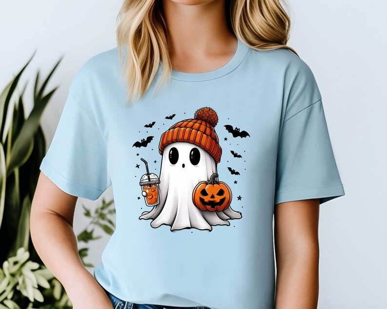 May include: A light blue t-shirt featuring a cartoon ghost wearing a knitted hat and holding a pumpkin and a cup of orange liquid. The ghost is surrounded by bats and stars.