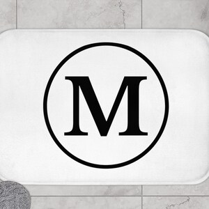 Puede incluir: Alfombra de baño blanca con un círculo negro que contiene la letra "M" en negrita.