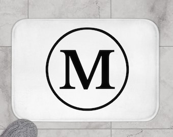 Puede incluir: Alfombra de baño blanca con un círculo negro que contiene la letra "M" en negrita.