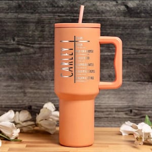 May include: A peach-colored tumbler with a handle and a straw. The tumbler features the name "CARLEY" vertically, a cross, and inspirational text. The design is in silver. The tumbler is on a wooden surface.