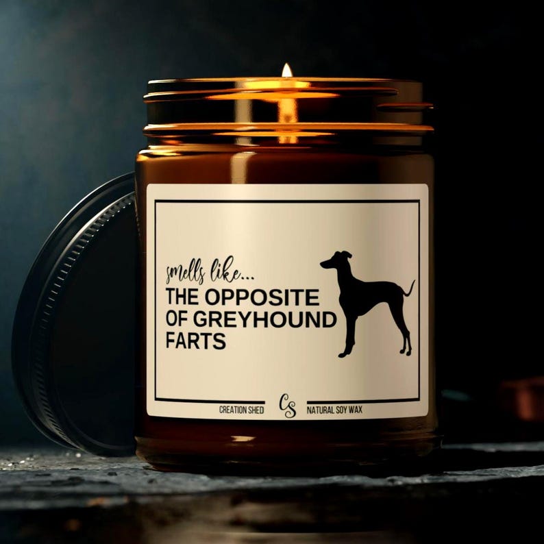 K&ouml;nnte beinhalten: Eine brennende, bernsteinfarbene Kerze in einem Glas mit schwarzem Deckel. Das Etikett tr&auml;gt die Aufschrift "smells like... THE OPPOSITE OF GREYHOUND FARTS" mit einer Windhund-Silhouette. Hergestellt aus nat&uuml;rlichem Sojawachs.