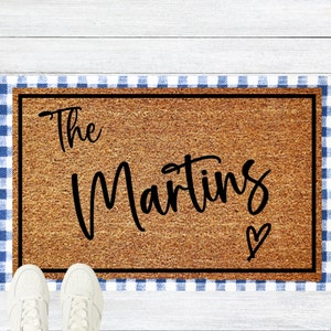 May include: A brown coir doormat with a black border and a blue and white checkered border. The text "The Martins" is written in black cursive lettering on the doormat.