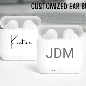May include: Two white wireless earbud cases with personalized names. The case on the left says "Kristina" and the case on the right says "JDM".
