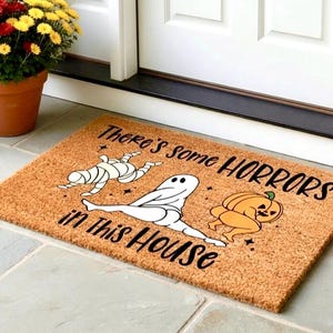 May include: A rectangular coir doormat with the text "There's some HORRORS in this HOUSE" in black. The mat features cartoon illustrations of a mummy, a ghost, and a pumpkin. The mat is brown and placed in front of a white door.