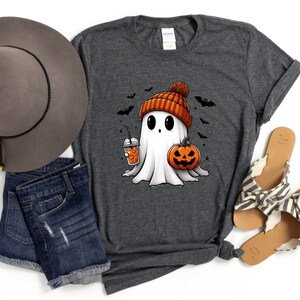 May include: A grey t-shirt featuring a cartoon ghost wearing an orange and white bobble hat, holding a pumpkin and a cup of orange liquid. The ghost is surrounded by bats.