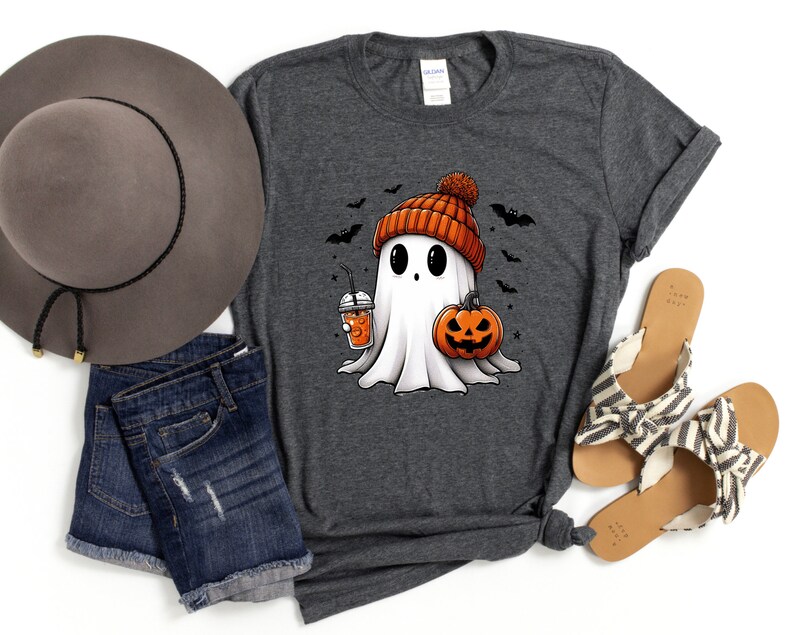 May include: A grey t-shirt featuring a cartoon ghost wearing an orange and white bobble hat, holding a pumpkin and a cup of orange liquid. The ghost is surrounded by bats.
