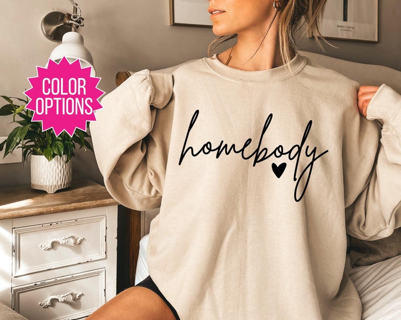 K&ouml;nnte beinhalten: Ein beiger Pullover mit dem Text "homebody" in schwarzer Schrift mit einem kleinen schwarzen Herzen.