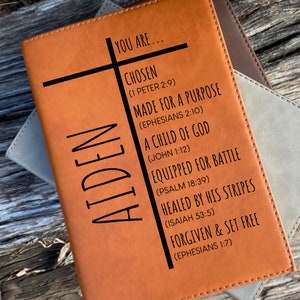 May include: A tan leather journal with the name "AIDEN" vertically printed on the left side. The journal features a black cross and inspirational text: "YOU ARE... CHOSEN, MADE FOR A PURPOSE, A CHILD OF GOD, EQUIPPED FOR BATTLE, HEALED BY HIS STRIPES, FORGIVEN & SET FREE."