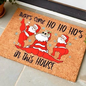May include: A rectangular doormat made of natural fibers. It features a humorous design of three Santas in red suits with white beards and sunglasses. The text reads "There's some HO HO HO's in this HOUSE."