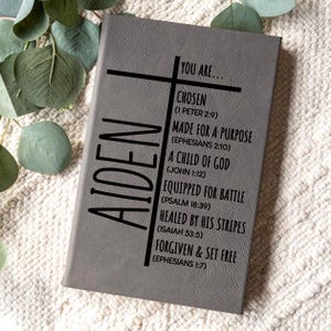 May include: Gray journal with the name "AIDEN" vertically on the left side. The journal features a cross design and inspirational text including "YOU ARE... CHOSEN" and "MADE FOR A PURPOSE". The journal is on a white textured surface.
